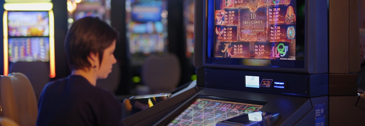 A person is seated at a slot machine console in a casino, focusing intently on the game displayed on the screen. The background shows several other illuminated gaming machines, adding a vibrant and colorful atmosphere to the scene.